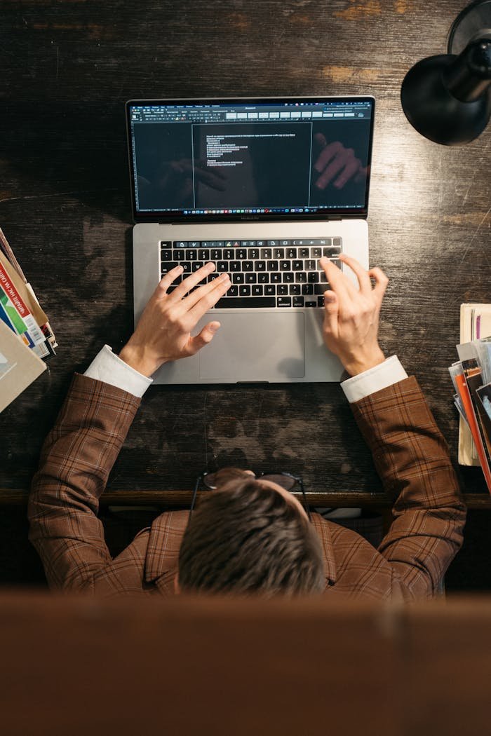 A person in a suit types on a laptop keyboard at a wooden desk, viewed from above.